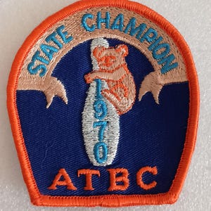 May include: A vintage patch with a blue background and an orange border. The patch features the text "STATE CHAMPION" in light blue, arched across the top. A koala bear clings to a bowling pin with the year "1970" in blue. The letters "ATBC" are at the bottom.