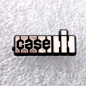 May include: A small, rectangular metal emblem with the word "Case" in a stylized font. The letters are in a light color, possibly silver or white, set against a black background. The emblem has a small, raised rectangular section on one end.