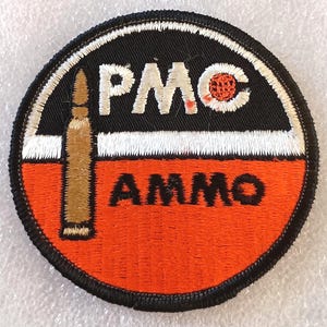 May include: A circular patch with a black border, featuring the text "PMC AMMO" in white and orange. A brown bullet is vertically positioned on the left. The background is black and orange.