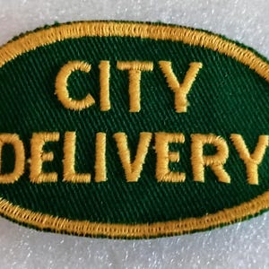 May include: An embroidered patch with the words "CITY DELIVERY" in gold thread on a dark green oval. The oval is outlined with a gold border, creating a classic and recognizable design.