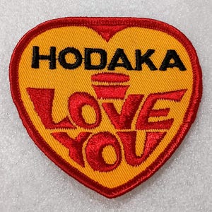 May include: Embroidered patch in the shape of a heart, featuring the word "HODAKA" above "LOVE YOU" in red lettering on a yellow background, with a red border.