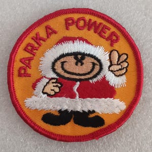 May include: Embroidered patch with the words "PARKA POWER" in red on a yellow background. The patch features a cartoon character in a red parka with white trim, giving a peace sign. The patch has a red border.