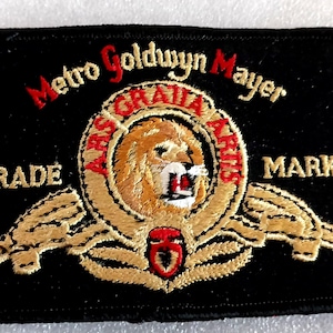 May include: Black rectangular patch with the Metro Goldwyn Mayer logo. The logo features a roaring lion inside a gold wreath, with the words "ARS GRATIA ARTIS" and "TRADE MARK" in red and white.