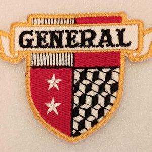 May include: Embroidered patch featuring a shield design with the word "GENERAL" in black on a white banner. The shield is red and black with white stars and a geometric pattern, outlined in gold.