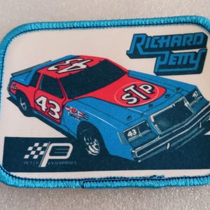 May include: Rectangular patch featuring a race car with the number 43, in blue, red, and white. The car has the STP logo on the hood and the words "Richard Petty" above it. The patch has a blue border and the Petty Enterprises logo.