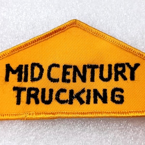 May include: A yellow, triangular patch with the words "MID CENTURY TRUCKING" embroidered in black. The patch has a stitched border and a retro aesthetic.