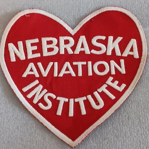 May include: A heart-shaped patch with a red background and white text. The text reads "NEBRASKA AVIATION INSTITUTE" in a bold, sans-serif font. The patch has a white border.