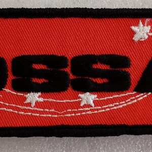 May include: Embroidered rectangular patch with the word "OSSA" in black block letters on a red background. The patch has a black border and white star and flower designs.