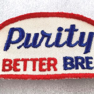 May include: Embroidered patch with a white background, red border, and the words "Purity is Better Bread" in blue and red lettering. The patch has a curved top and a rectangular bottom.