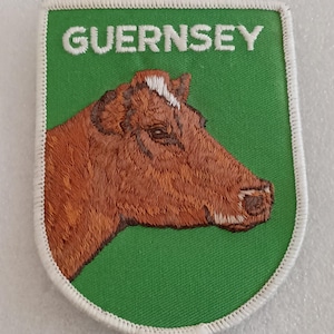 May include: Embroidered patch featuring a brown cow's head on a green background. The word "GUERNSEY" is embroidered in white at the top. The patch has a white border.
