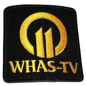 May include: A rectangular black embroidered patch featuring a gold circular logo with the number 11 inside. Below the circular emblem, the text WHAS-TV is embroidered in gold thread. The patch has a black stitched border around the entire edge.