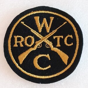 May include: A round, black patch with gold embroidered text and design. The text reads "W RO TC C" around the perimeter. Two crossed rifles are in the center. The patch has a gold border.