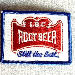 May include: A rectangular patch with a white background and a blue border. The patch features the words "L.B.C. ROOT BEER" in red, with the phrase "Still the Best" in blue. The design is reminiscent of vintage advertising.