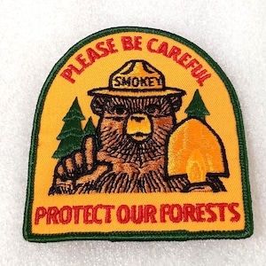 May include: Embroidered patch featuring Smokey Bear in a ranger hat, with the text "PLEASE BE CAREFUL" and "PROTECT OUR FORESTS". The patch has a yellow background with green and brown details.