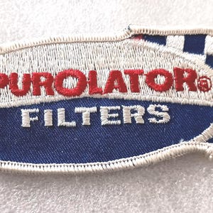 May include: Embroidered patch with the text "PUROLATOR FILTERS" in red and white. The patch has a blue and white checkered flag design on the right side. The patch is oval-shaped with a white border.