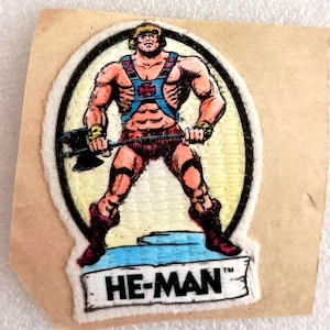 May include: Vintage He-Man patch featuring a muscular, cartoon character with blonde hair, wearing blue and red armor, and holding an axe. The patch is in an oval shape with a black border and the text "HE-MAN" at the bottom.
