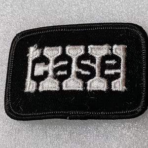 May include: Black rectangular patch with the word "CASE" embroidered in white block letters. The patch has a black border and is likely an emblem or logo.