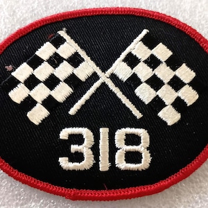 May include: An oval-shaped embroidered patch with a black background and a red stitched border. The design features two crossed white and black checkered racing flags positioned above the white embroidered numbers 318 centered at the bottom.