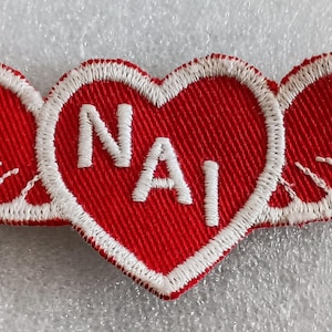 May include: A red and white embroidered patch featuring a heart with the letters "NAI" inside, flanked by wings. The wings and heart are outlined in white stitching, creating a striking contrast against the red fabric. This patch is likely a decorative emblem.