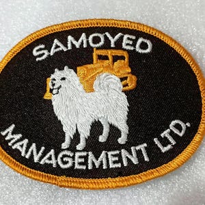 May include: Embroidered patch with a black oval background and a gold border. The patch features a white Samoyed dog and a gold tractor. The text "SAMOYED MANAGEMENT LTD." is in white.