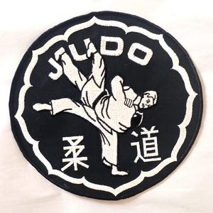 May include: A circular black embroidered patch featuring a white border with a scalloped edge. The patch displays a white graphic of two figures performing a judo throw, the word JUDO in white letters, and two Japanese kanji characters in white.