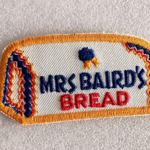 May include: Embroidered patch featuring the text "MRS BAIRD'S BREAD" in navy blue and red, with a blue and gold emblem. The patch has an orange border and a white background with a textured pattern.