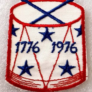 May include: Embroidered patch featuring a drum design with red and blue accents. The patch has a white background with blue stars and the numbers "1776 1976" in blue. Two blue drumsticks are crossed at the top.