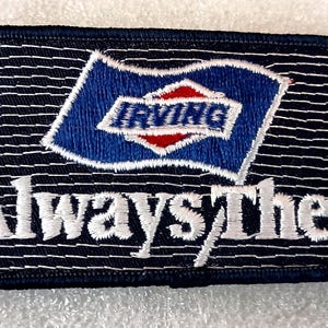 May include: A rectangular embroidered patch with a dark blue background and thin horizontal white lines. It features a blue flag graphic with a red and white diamond shape containing the text IRVING, and the white text Always There below.