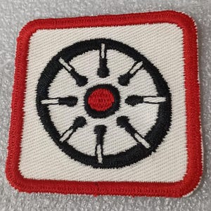 May include: Square patch with a white background, featuring a black wheel design with a red center. The patch has a red border. The wheel design has eight spokes and a central red circle.