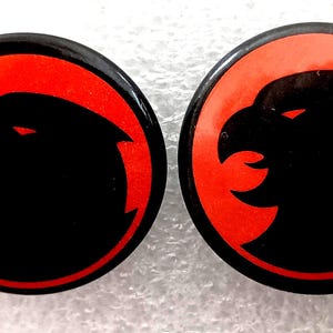 May include: Two round, black buttons with a red border and a black eagle silhouette. The eagle has a sharp beak and is facing left. The buttons are glossy and appear to be made of plastic or metal.