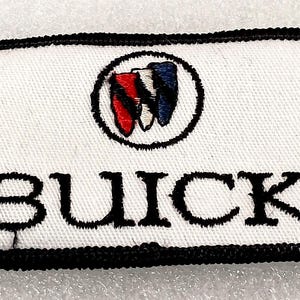 May include: Rectangular white patch with a black border featuring the word "BUICK" in black block letters. Above the text is a circular emblem with red, white, and blue stylized shield shapes.