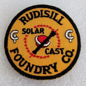 May include: Round yellow patch with black and white text and graphics. The patch reads "RUDISILL SOLAR FOUNDRY CAST" in a circular pattern. A red and white graphic of a planet is in the center, crossed out with a black line. Two white "G" shapes are on either side.
