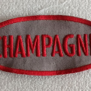 May include: An oval-shaped patch with the word "CHAMPAGNE" embroidered in red on a gray background. The patch has a red border. The fabric appears to be a woven material.