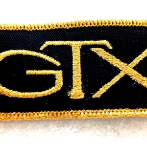 May include: A rectangular embroidered patch featuring a black fabric background with the letters GTX stitched in gold thread. The patch is finished with a gold stitched border around the entire perimeter of the rectangular shape.