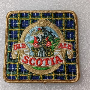 May include: Embroidered patch with a square shape and a gold border. The background features a blue and green plaid pattern. The center design includes a banner with the words "OLD ALE SCOTIA" and a detailed illustration of a landscape.
