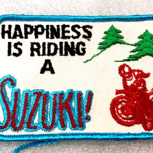 May include: Embroidered patch with the text "HAPPINESS IS RIDING A SUZUKI!" in black and red. Features a red motorcycle graphic, green trees, and a blue border. A motorcycle-themed item.