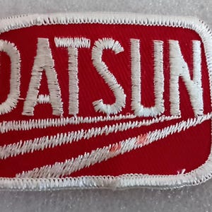May include: Embroidered patch with the word "DATSUN" in white, stitched lettering on a red rectangular background. The patch has a white stitched border and diagonal white lines.