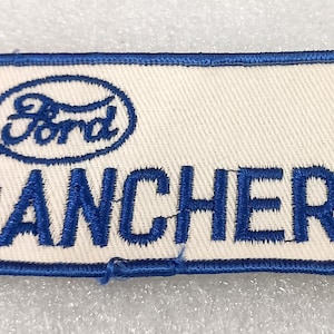 May include: Rectangular patch with a white background and a blue border. The Ford logo is at the top, with the word "RANCHERO" in bold blue letters below. The patch is likely for a Ford Ranchero vehicle.