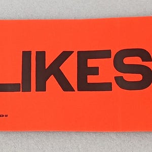 May include: An orange rectangular sign with the text "USA LIKES LBJ" in black lettering. The sign appears to be a vintage political statement.
