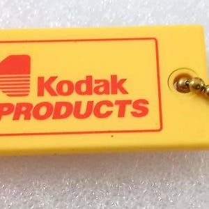 May include: Yellow Kodak PRODUCTS whistle with a red logo and text. The whistle has a small hole for a ball chain. The ball chain is gold-colored.