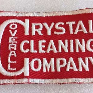 May include: A rectangular red patch with white embroidered text. The text reads "OVERALL CRYSTAL CLEANING COMPANY". The patch has a white border and is likely an emblem or logo.