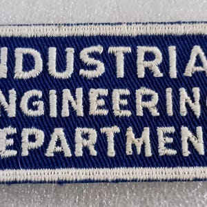 May include: A rectangular patch with the words "INDUSTRIAL ENGINEERING DEPARTMENT" embroidered in white on a blue background. The patch has a white border.
