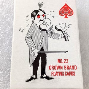 May include: A white playing card box with a cartoon illustration of a man playing a broken violin. The box features the text "NO.23 CROWN BRAND PLAYING CARDS" in red. A red spade symbol is in the upper right corner.
