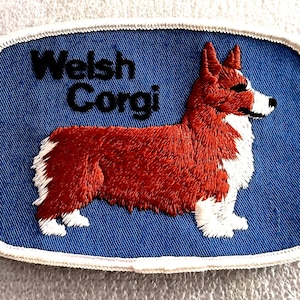 May include: Embroidered patch featuring a Welsh Corgi dog. The patch has a blue background with the words "Welsh Corgi" in black. The dog is depicted in red and white, with detailed stitching and a white border.