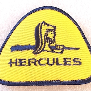 May include: Yellow triangular patch with a blue border and a blue image of a figure holding a weapon. The word "HERCULES" is embroidered in blue across the bottom of the patch.