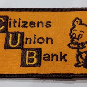 May include: Embroidered patch with the text "Citizens Union Bank" and a cartoon bear. The patch is rectangular with rounded corners, a brown border, and a gold background. The text and bear are brown.