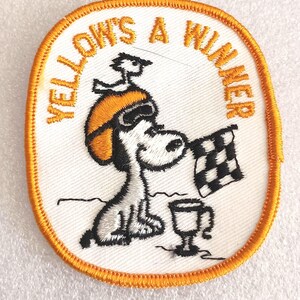 May include: Embroidered patch featuring Snoopy in a racing helmet, holding a checkered flag and a trophy. The patch has an oval shape with an orange border and the text "YELLOW'S A WINNER" in orange.