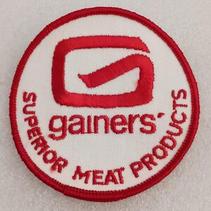 May include: A circular patch with a white background and a red border. The patch features the word "gainers" in red, with a stylized red "G" logo above it. The words "SUPERIOR MEAT PRODUCTS" are also in red, encircling the design.