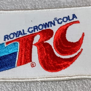 May include: Embroidered patch featuring the Royal Crown Cola logo. The patch has a white background with the words "Royal Crown Cola" in blue and the letters "RC" in red. A blue and turquoise stripe is on the left.