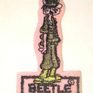 May include: Embroidered patch featuring a cartoon character in a long coat and hat. The character is in shades of green and gold, with black outlines, on a pink background. The word "BEETLE" is embroidered at the bottom.